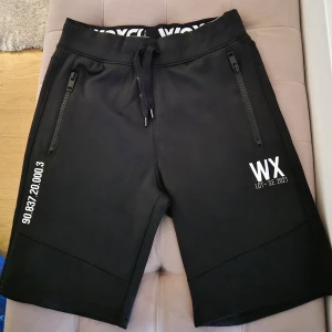 Svarta shorts från WOXO 720° - Svarta shorts från WOXO 720° med tryckta vita detaljer och logga. De har elastisk midja med snörning, två dragkedjeförsedda fickor fram och urban streetwear-vibe. Perfekta för chill dagar eller träning. Materialet är mjukt och stretchigt för extra komfort.