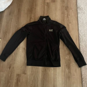 Svart EA7 träningsset med dragkedja - Säljer ett svart träningsset från EA7 Emporio Armani. Setet består av en zip-jacka med hög krage och matchande byxor, båda med EA7-logga på bröst och lår. Materialet är mjukt och stretchigt, perfekt för sport eller chill. Jackan har dragkedja och fickor.
