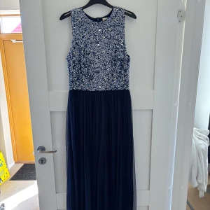 Mörkblå långklänning Lace & Beads - Supersnygg långklänning från Lace & Beads i mörkblått med glittrig paljettöverdel och pärldetaljer. Ärmlös modell med rund halsringning och flowy kjol i tunt tyg. Perfekt för bal eller festliga tillfällen!