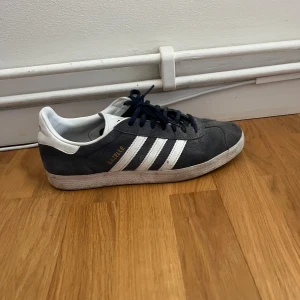 Adidas Gazelle sneakers i grå mocka - Klassiska Adidas Gazelle sneakers i mörkgrå mocka med vita detaljer och de ikoniska tre ränderna på sidan. Skon har snörning och låg profil, perfekt för en avslappnad streetstyle-look.