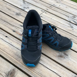 Svarta och blå Salomon sneakers strl 33 - Säljer ett par svarta och blå sneakers från Salomon i storlek 33. Skorna har snabbsnörning, grov mönstrad sula för extra grepp och är tillverkade i syntetmaterial och mesh. Perfekta för dig som gillar att vara aktiv utomhus.