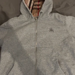 Grå hoodie från Burberry med dragkedja - Säljer en grå hoodie från Burberry med klassiskt rutigt foder i huvan. Tröjan har dragkedja framtill, huva med snörning och broderad logga på bröstet. Perfekt för dig som gillar stilrena och exklusiva plagg med en twist.