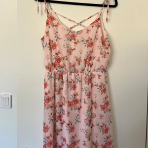 Ljusrosa blommig sommarklänning - Supersöt ljusrosa klänning med röda och rosa blommor, smala axelband med knyt och korsad detalj i ryggen. 