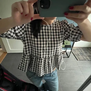 Rutig blus med volangkant - Supersöt blus i svartvitt rutigt mönster med volangkant nedtill. Modellen har rund hals och korta ärmar med lätt puff. 
