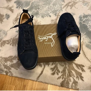 Mörkblå sneakers från Christian Louboutin - Snygga mörkblå sneakers från Christian Louboutin i mocka med klassiska röda sulor och diskret broderad logga på hälen. Skorna har rund tå, snörning och platt sula. Perfekta för dig som vill ha en lyxig och stilren look med en touch av attityd.