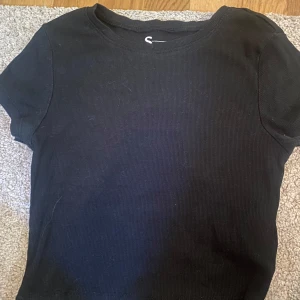 Svart croppad topp från FSBN Sister - En svart, ribbad croppad topp från FSBN Sister. T-shirten har rund halsringning och korta ärmar. Enkel och stilren modell som är lätt att matcha till olika outfits. Perfekt för dig som gillar en basic men trendig look.