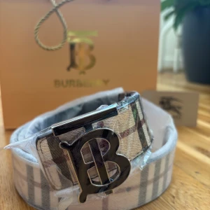 Burberry rutigt skärp med B-spänne - Snyggt Burberry-bälte i klassiskt rutigt mönster med stora B-spännet i metall. Ena sidan är beige med svarta och röda rutor, andra sidan är helsvart med diskret rutmönster och Burberry-ryttare. Tillverkat i skinn och levereras med originalpåse och box. Storlek fixare ingår!