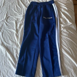 Blåa Palm Angels Byxor - Snygga blåa mjukisbyxor från Palm Angels med vit logga på framsidan och vita ränder längs sidorna. Byxorna har elastisk midja och raka ben för en chill och trendig look. Byxorna är i ett jättebra skick!