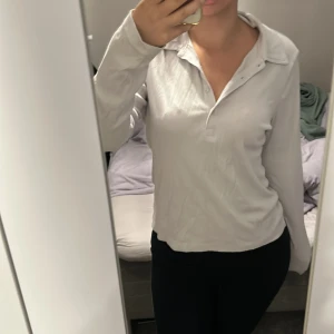 Vit långärmad pikétröja i bomull - Säljer en stilren vit pikétröja med lång ärm och klassisk krage. Tröjan har knappar framtill och är gjord i mjuk bomull med diskret ribbad struktur. Perfekt för en clean och enkel look. Passar dig som gillar basic men ändå vill ha något med lite edge.