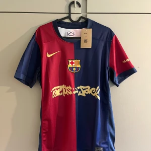 FC Barcelona Nike fotbollströja S - FC Barcelona matchtröja från Nike i Dri-FIT-material. Halva tröjan är vinröd och andra halvan marinblå, med klubbmärke och Nike-logga i guld. Tryck på ärmen och coolt grafittiliknande tryck på magen. Klassisk rund hals och korta ärmar.