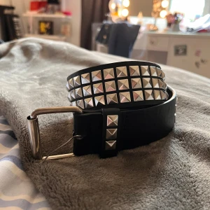 Svart nitarbälte - Säljer ett svart bälte med tre rader fyrkantiga silverfärgade nitar och silverspänne. Bältet är brett och har metallhål för justering. Perfekt accessoar för en edgy look och passar till jeans eller kjol. Den har små defekter men fungerar som den ska! Asball och skön💕 Den är ungefär 119cm lång med minsta möjliga hål placering vid 94,5 ish.