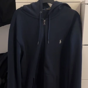 Marinblå hoodie från Ralph Lauren - Snygg marinblå hoodie från Ralph Lauren med dragkedja framtill och klassisk vit logga på bröstet. Tröjan har huva med snörning, känguruficka och långa ärmar. Perfekt för en chill och stilren look.