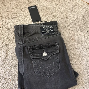 Svarta jeans från True Religion(Joey low rise flare) - Snygga svarta jeans från True Religion med klassiska kontrastsömmar och stora fickor bak med lock och silverfärgade knappar. Jeansen har en rak passform och normal midja. Materialet är mjukt denim som sitter skönt på kroppen. Helt oanvända, säljs på grund av för stor storlek.(Pris går att diskutera)