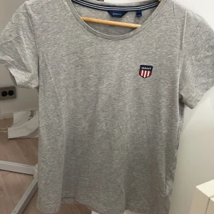 Grå t-shirt från GANT med logga - Snygg gråmelerad t-shirt från GANT med klassisk rund hals och korta ärmar. På bröstet sitter en broderad GANT-logga i form av en sköld med rödvita ränder. T-shirten är i mjuk bomull och har en clean, stilren look.