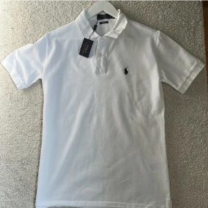 Polo Ralph Lauren  - Klassisk vit pikétröja från Polo Ralph Lauren i bomull med broderad logga på bröstet. Tröjan har korta ärmar, krage och knäppning med två knappar framtill. Perfekt för en clean och stilren look.