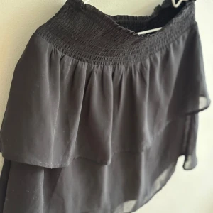 Svart volangkjol från ONLY - Svart kortkjol från ONLY med bred smock i midjan och dubbla volanger. Kjolen är luftig och har en flowy känsla, perfekt för dig som gillar en enkel men ändå trendig look.