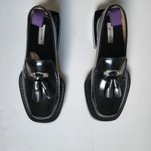 Svarta loafers med tofs från Eytys/ Eytys Rio - Unika svarta loafers från Eytys i glansigt skinn med fyrkantig tå, chunky klack och dekorativa tofsar framtill. Snygga detaljer som metallspänne och lila hälflik. Perfekta för dig som vill sticka ut med en edgy och modern look.