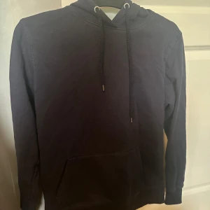 Mörkblå hoodie med huva och ficka - En stilren mörkblå hoodie med huva och dragsnören, samt en stor magficka framtill. Perfekt för chill dagar eller när du vill ha en enkel och bekväm look. Långärmad och i mjukt bomullsmaterial.