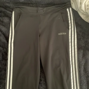 Svarta Adidas mjukisbyxor med ränder - Svarta mjukisbyxor från Adidas med klassiska vita ränder längs sidorna och Adidas-logga på låret. Byxorna har fickor och resår i midjan med plats för snörning. Perfekta för chill eller träning. Obs: inget snöre i