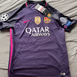 FC Barcelona Suárez 9 Champions League tröja - Snygg FC Barcelona matchtröja från Nike med Suárez 9 på ryggen. Tröjan är mörklila med svarta ärmar och rosa detaljer, samt tryck från Champions League och Qatar Airways. Broderade klubbmärken och Dri-FIT-material som andas. Perfekt för dig som älskar Barca!