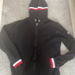 Svart hoodiejacka från Moncler - Svart hoodiejacka från Moncler med hel dragkedja, huva och ribbade muddar i rött, vitt och grått. Jackan har fickor framtill och Moncler-logga på ärmen. Perfekt för dig som gillar stilren streetwear med snygga detaljer.