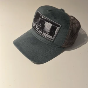 Gråblå truckerkeps från emilio pendenza - Snygg truckerkeps i gråblått med mesh baktill och justerbar snapback. Framsidan har en svartvit patch med coolt fotomotiv. Kepsen har mjuk, nästan sammet-liknande känsla framtill och klassisk böjd skärm. Pris ej huggt i sten!! Mvh