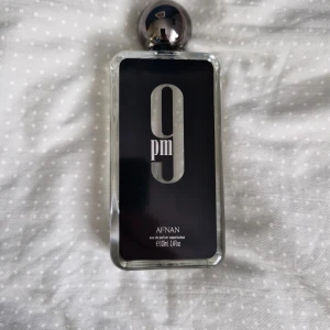 Afnan 9pm Eau de Parfum 100ml - Afnan 9pm Eau de Parfum i en stilren, rektangulär glasflaska med svart etikett och silverdetaljer. Flaskan rymmer 100 ml och har en metallisk, rund kork. Parfymen levereras i en svart, räfflad kartong med silvertext. Doften är känd för sina varma och kryddiga toner. Den är fylld till 85%
