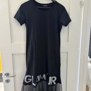 Svart t-shirtklänning från Angie - Svart t-shirtklänning från Angie med korta ärmar och rund hals. Nederdelen har en transparent meshvolang. Skön och avslappnad passform