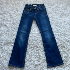 Blå bootcut jeans Gina Tricot 152 - Snygga blå jeans från Gina Tricot i storlek 152. Modellen har bootcut-ben, hög midja och klassiska fickor fram och bak. Jeansen har dubbla silverknappar och kontrastsömmar för en cool look. Perfekta till sneakers eller boots.