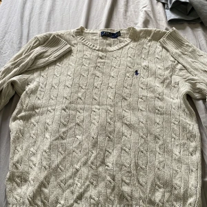 Vit kabelstickad tröja Polo Ralph Lauren - Kabelstickad vit tröja från Polo Ralph Lauren med klassisk rund halsringning och broderad marinblå logga på bröstet. Tröjan är i bomull och har långa ärmar med ribbade muddar. Perfekt för dig som gillar en clean och tidlös stil.