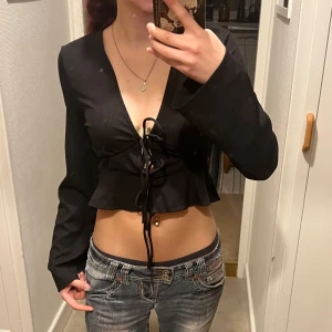 Svart ribbad topp med knytning - Säljer en svart ribbad topp med långa ärmar och knytning framtill. Toppen är croppad och har en v-ringad design som ger en stilren look. Perfekt för en trendig outfit!