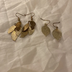 Guldiga lövformade örhängen - Två par guldiga örhängen i form av löv. Det ena paret har flera små löv i lager, medan det andra paret har ett enkelt löv. Perfekta för att ge en naturlig touch till din stil.