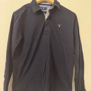 Mörkblå pikétröja från GANT - Säljer en stilren mörkblå pikétröja från GANT med broderad logga på bröstet. Tröjan har långa ärmar och en klassisk krage med knappar. Perfekt för en avslappnad men ändå snygg look.