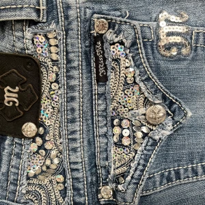Blå Miss Me jeans med paljettdetaljer  - Snygga Miss Me jeans med unika paljettdetaljer på fickorna. De har en bootcut-stil och är dekorerade med silverfärgade knappar. Perfekta för att ge en extra touch till din outfit. Det finns ett litet hål vid knappen men det är inget som försämrar användningen 
