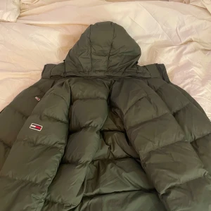 Pufferjacka från Tommy Hilfiger - Snygg grön dunjacka från Tommy Hilfiger med huva och dragkedja. Jackan har en quiltad design och märkeslogga på ärmen. Perfekt för kyliga dagar.