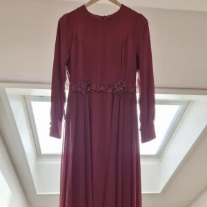 Vinröd aftonklänning med pärlor - Elegant vinröd aftonklänning med långa ärmar och vacker pärldetalj i midjan. Perfekt för en festlig kväll eller speciellt tillfälle. Klänningen har en klassisk rund hals och ett flytande fall som ger en sofistikerad look.