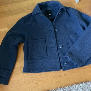 Mörkblå jacka från 157 - Säljer en stilren mörkblå jacka från lager 157. Jackan har en klassisk design med stora knappar och två praktiska fickor framtill. Perfekt för kyligare dagar och ger en snygg look. Passar bra till både jeans och chinos.