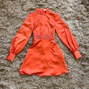 Orange satinklänning med öppen rygg - Orange satinklänning med långa ärmar och med band i ryggen man kan knyta. Ryggen är öppen. Använda ett fåtal gånger