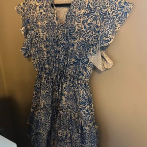 Blå och vit klänning från Hollies - Säljer en vacker blå och vit klänning från Hollies med ett detaljerat mönster. Klänningen har korta ärmar och en v-ringad design.🥂💋