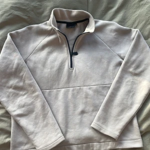 Grå tröja från Sail Racing - Säljer en stilren beige tröja från Sail Racing med halv dragkedja framtill. Tröjan har långa ärmar och en hög krage, perfekt för kyligare dagar. Den är både bekväm och snygg, idealisk för en avslappnad look.