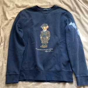 Ralph Lauren sweater - Säljer en snygg mörkblå sweatshirt från Ralph Lauren med en ikonisk björntryck på framsidan. Tröjan har långa ärmar och en rund halsringning. Perfekt för en avslappnad stil. 🐻