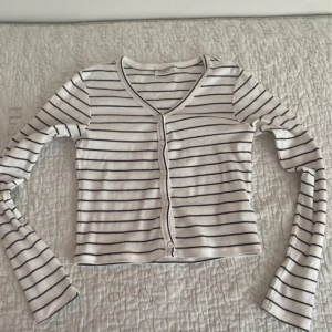 Randig vit och svart kofta från Kappahl - Säljer en snygg randig kofta från Kappahl i vitt och svart. Den har en v-ringad design med knappar framtill och långa ärmar. Perfekt för en stilren look!