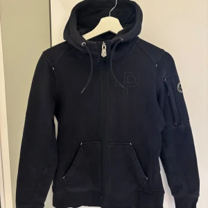Marinblå hoodie från Pelle P - Säljer en marinblå hoodie från Pelle P med dragkedja och huva. Tröjan har långa ärmar och en logotyp på bröstet samt en på ärmen. Perfekt för en casual look.