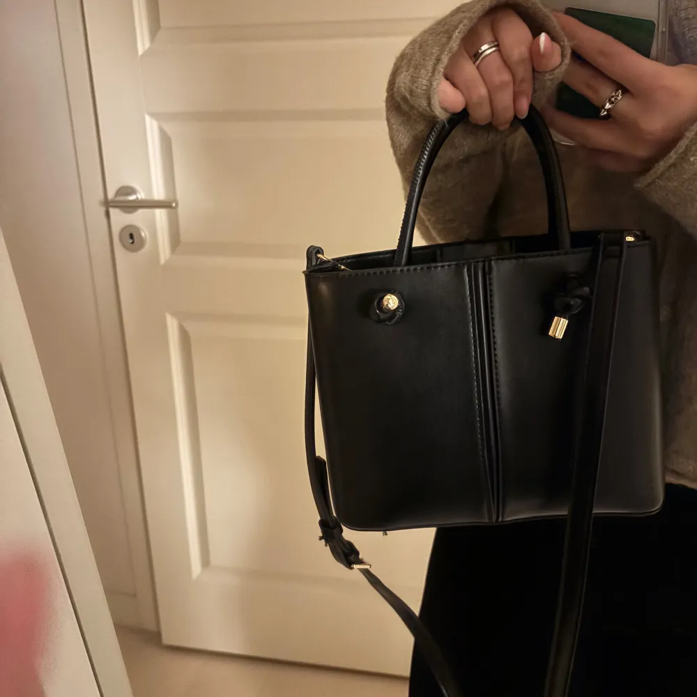 Snygg svart handväska i skinn med praktisk dragkedja och eleganta gulddetaljer. Väskan har en enkel och stilren design med två handtag för enkel bärning. Väskan är i väldigt bra skick.. Laukut & Käsilaukut.