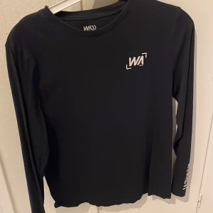Svart långärmad tröja från WA - Snygg svart långärmad tröja från WA med vit logga på bröstet och ärmen. Perfekt för en stilren och sportig look. Tröjan är gjord i ett mjukt och bekvämt material.