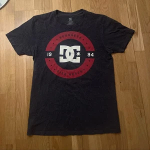 Mörkgrå t-shirt från DC Shoes - Säljer en mörkgrå t-shirt från DC Shoes med ett stort tryck i rött och vitt på framsidan. T-shirten har korta ärmar och en rund halsringning. 