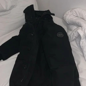 Svart dunjacka från Canada Goose - Säljer en svart dunjacka från Canada Goose. Jackan har en stilren design med knappar och en hög krage. Perfekt för kalla dagar med sin varma vaddering. Den har långa ärmar och en diskret logga på bröstet.
