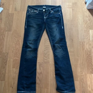 Mörkblå jeans från True Religion - Snygga mörkblå jeans från True Religion med kontrasterande vita sömmar och klassiska femficksdesign. Jeansen har en bootcut-stil och är dekorerade med knappar och broderade detaljer på bakfickorna. Innebenslängden 78,5cm, midjemått 40 cm