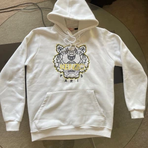Vit hoodie från Kenzo med tigerbroderi - Säljer en snygg vit hoodie från Kenzo med ett coolt tigerbroderi i gult och svart på framsidan. Hoodien har en klassisk känguruficka och justerbar huva med dragsko. Perfekt för en stilren look!