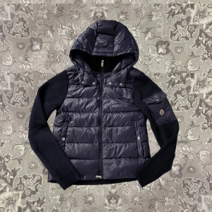 Svart pufferjacka från Moncler - Snygg svart pufferjacka från Moncler med huva och dragkedja. Jackan har långa ribbade ärmar och en quiltad kropp. Perfekt för kyligare dagar. Moncler-logga på ärmen ger en exklusiv touch.